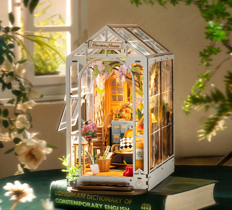 Miniature model of a garden house with interior details, placed on a book with plants around.